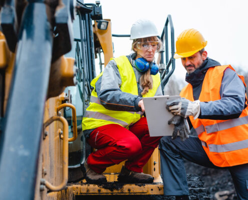 Deux machinistes de chantier en gilet haute visibilité analysent un plan sur tablette devant un engin de chantier.
