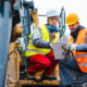 Deux machinistes de chantier en gilet haute visibilité analysent un plan sur tablette devant un engin de chantier.