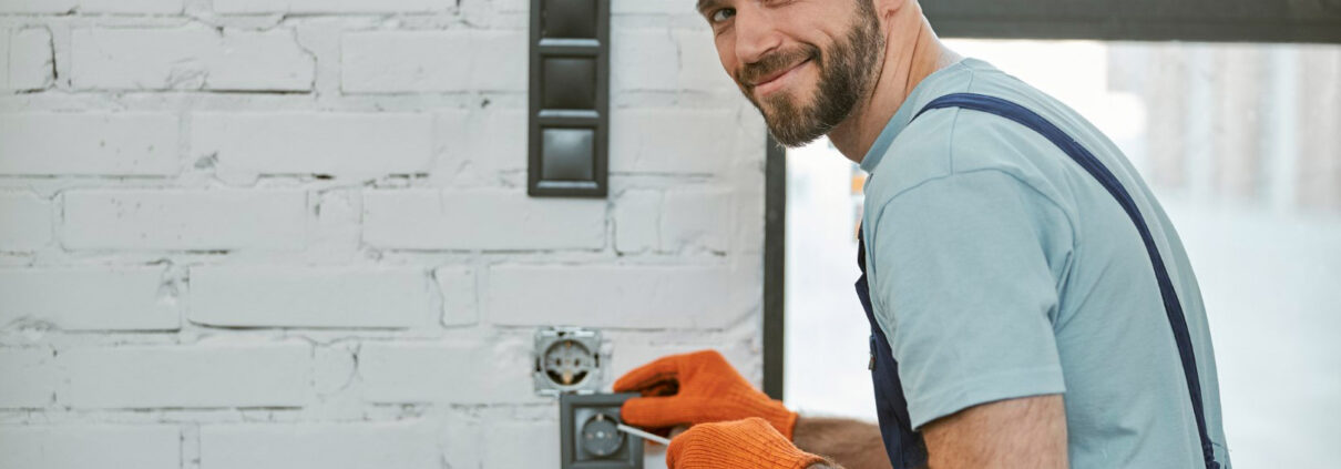 Monteur-électricien souriant installant une prise électrique murale dans un bâtiment.