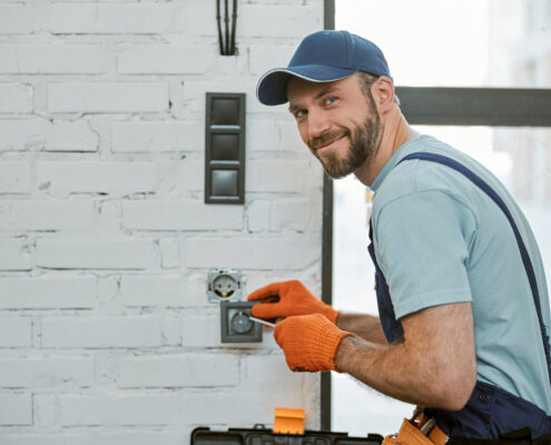 Monteur-électricien souriant installant une prise électrique murale dans un bâtiment.